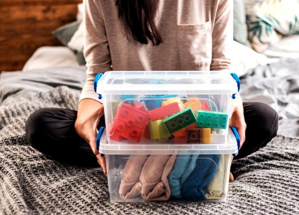 A woman sitting on a bed with clothes and toys sorted into plastic bins in front of her.