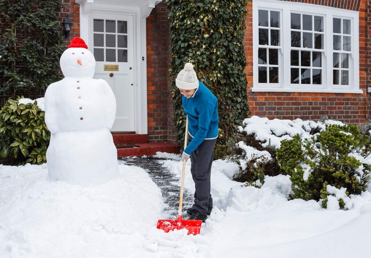 A person shoveling snow off a sidewalk near a snowman in front of a house during winter.