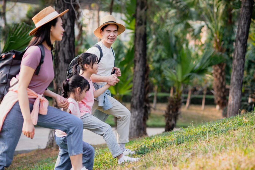 A smiling family with two kids hiking uphill in a sunny forest park, wearing casual clothes, hats, and backpacks.