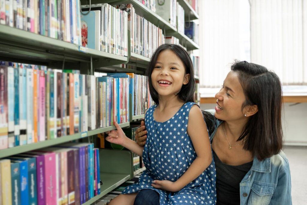 A smiling young girl in a library with her mother beside her.